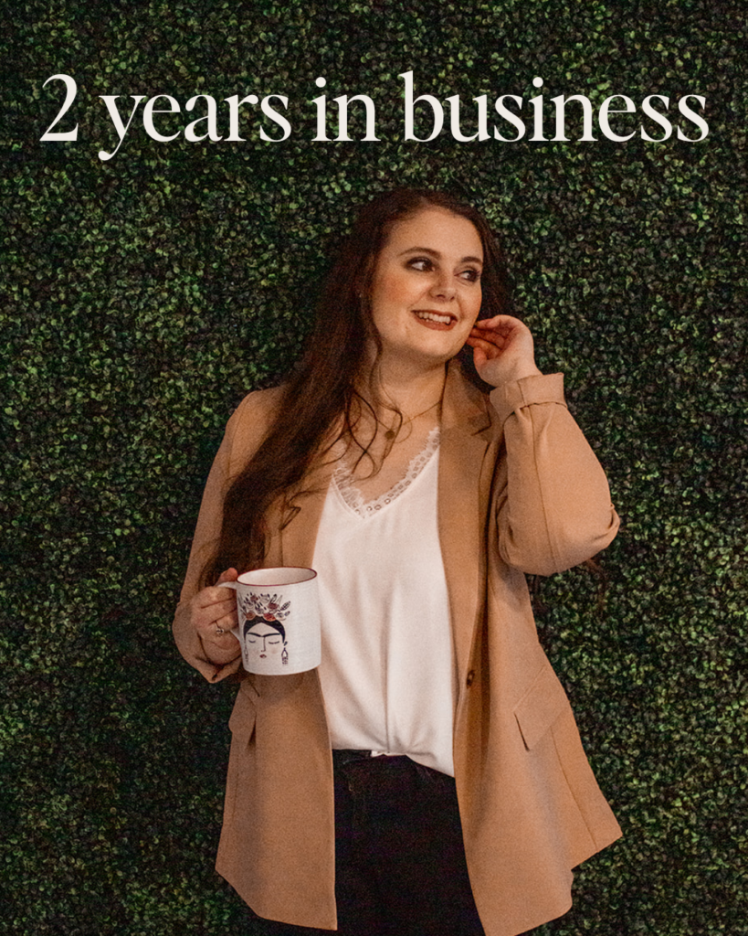 Emily Foster, owner of Emily Foster Creative, standing in front of an ivy wall and wearing a nude blazer. The text on image says "2 years in business"