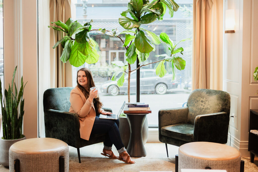 Emily Foster, owner of Emily Foster Creative, sipping coffee next to a big plant with a laptop on her lap