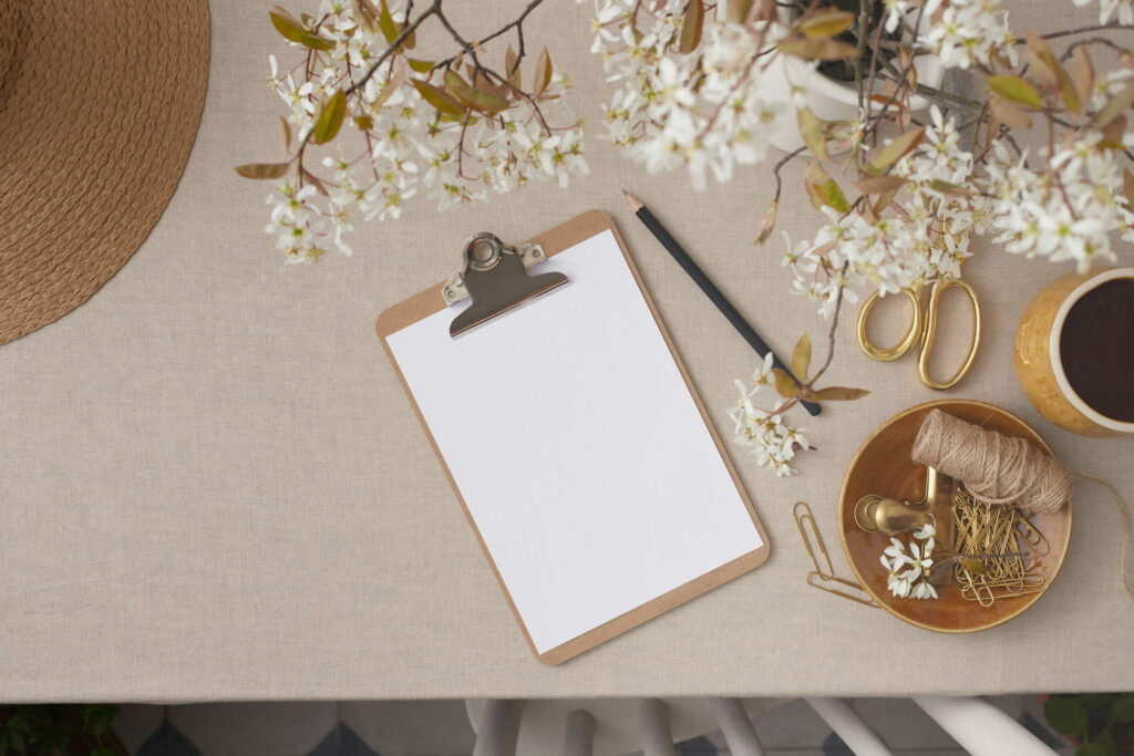 Flat lay of blank clipboard with white flowers, pencil, gold scissors, twine, and coffee on neutral fabric background.