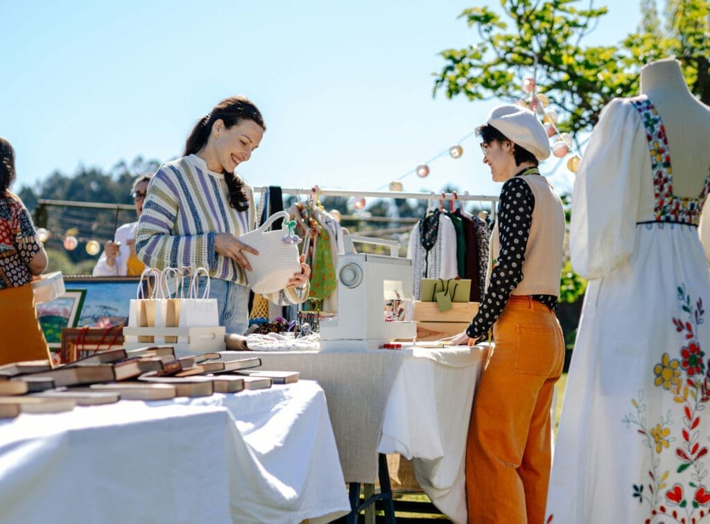 Two women chatting at an outdoor market with handmade items and clothing displays