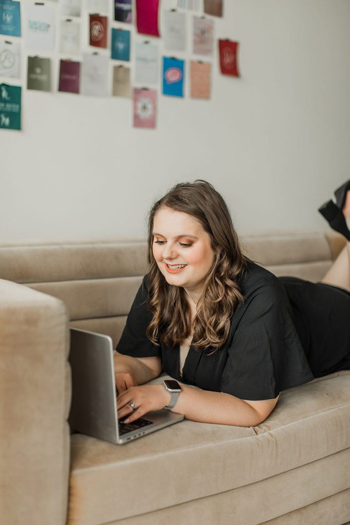 Woman wearing all black laying on a brown couch working on the computer and smiling.