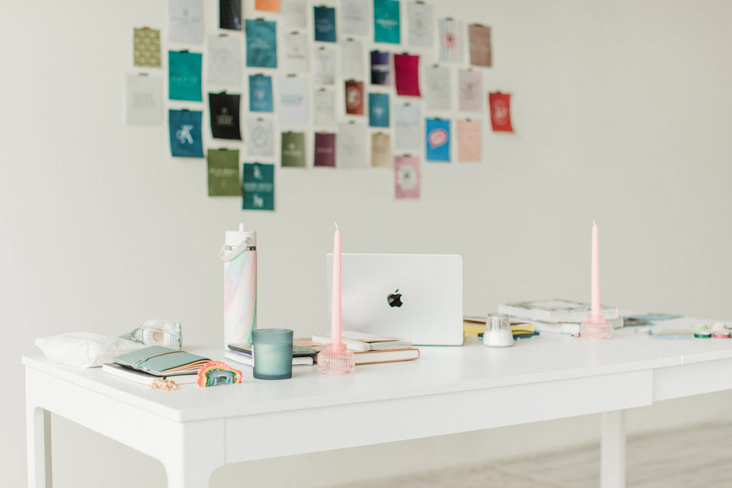 Wide shot of a desk with a computer, water bottle, candles, and notebooks on it. In the background is a mood board on the wall.