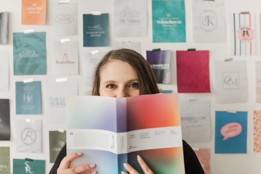Emily Foster holding a colorful design book in front of a wall of branding samples.